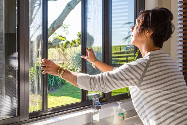 Two hands drawing pleated screens sideways across a kitchen window